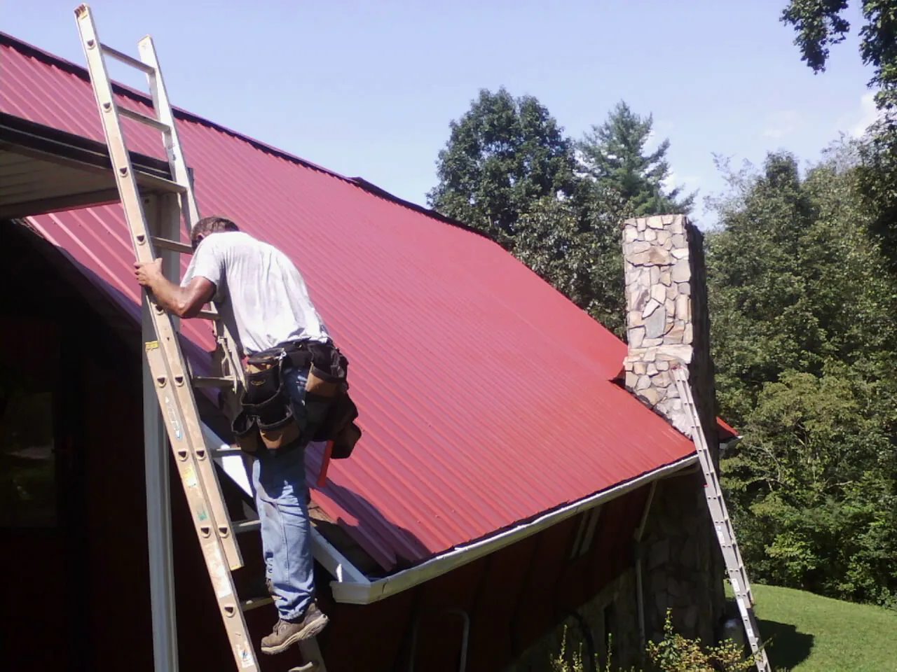 Roofer on a ladder beside a red metal roof and stone chimney — Soffit & Fascia Repair by Roofing Brothers in Celina